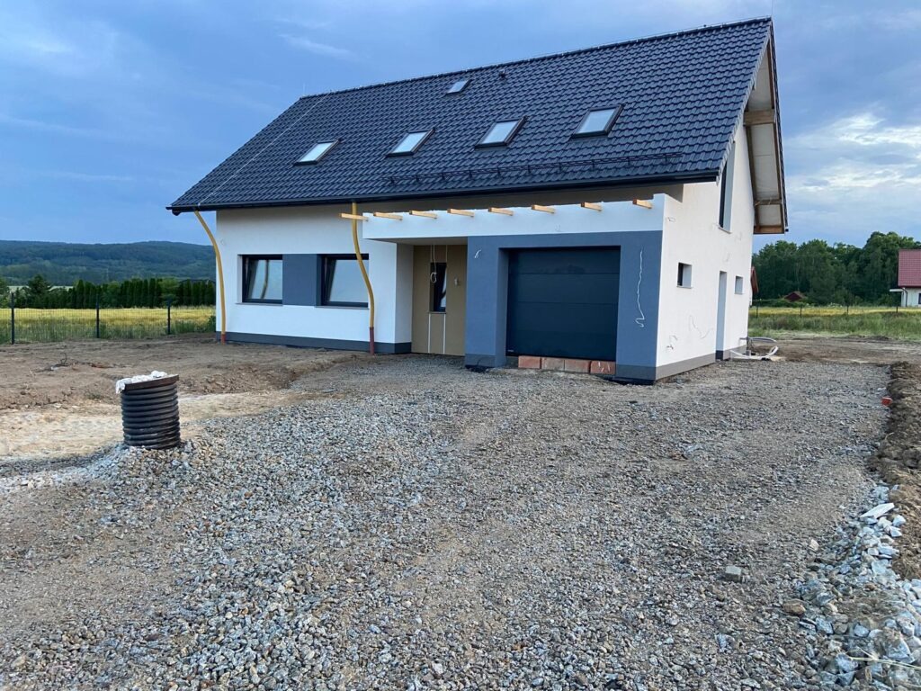 Nowoczesny dom jednorodzinny z garażem w trakcie wykończenia, widok z zewnątrz, szara dachówka, biała elewacja z szarymi akcentami, teren wokół domu przygotowany pod ogród.