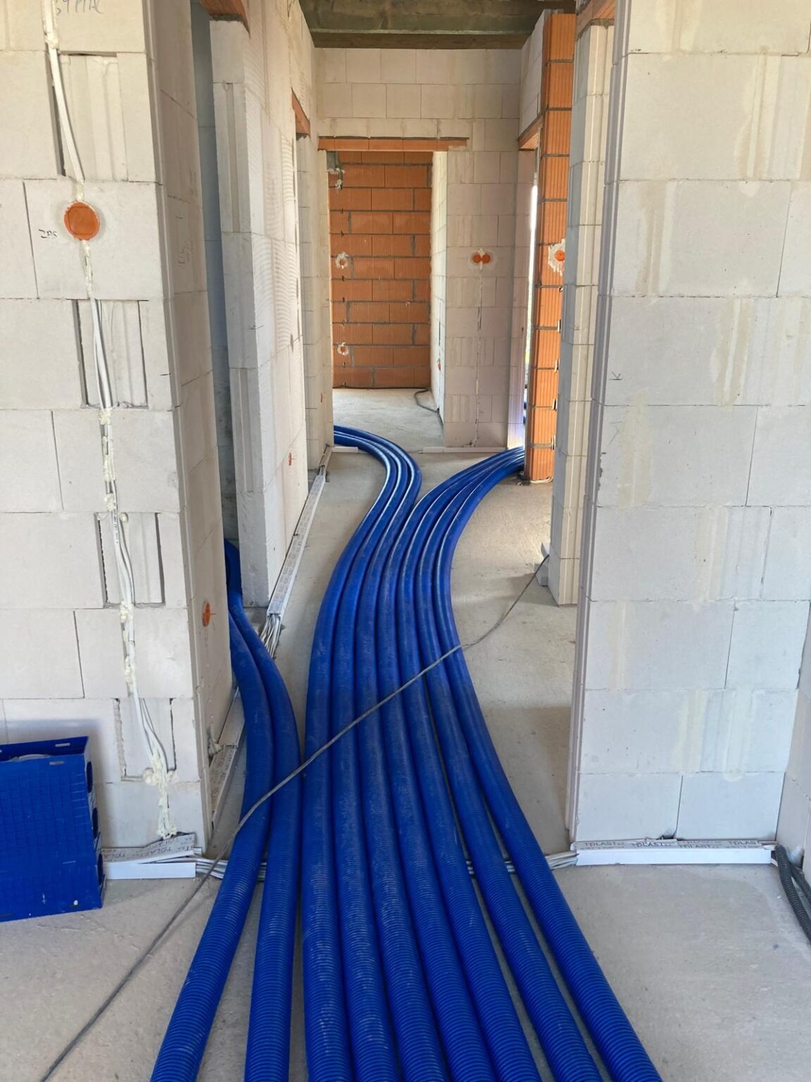 Wnętrze w trakcie budowy z widocznymi niebieskimi rurami instalacyjnymi na podłodze, ściany z białych bloczków, widoczne otwory drzwiowe.