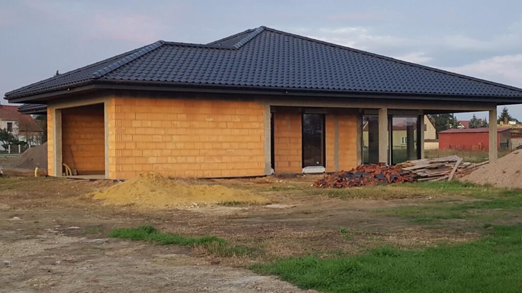 Niedokończony dom jednorodzinny z ciemną dachówką, widoczne ściany z pustaków i okna. Na pierwszym planie piasek i gruz. Budowa w toku, widok z zewnątrz.
