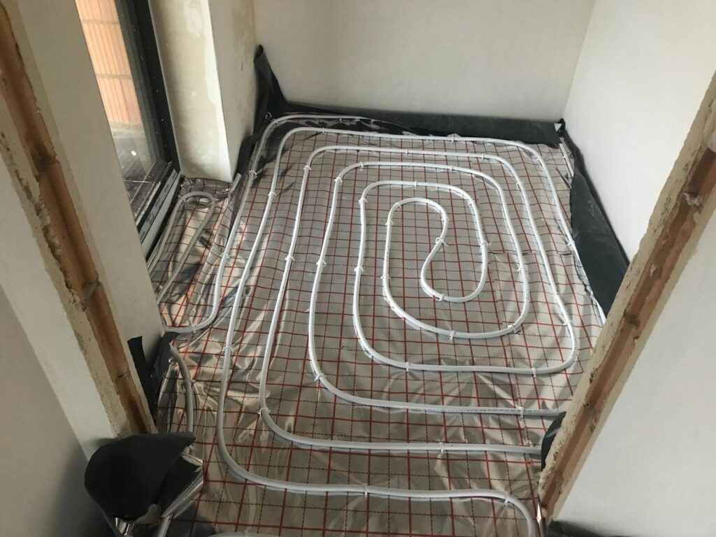 Białe rury ogrzewania podłogowego ułożone na folii z czerwoną kratką, widoczne fragmenty ścian w surowym stanie. Instalacja przed wylaniem posadzki.