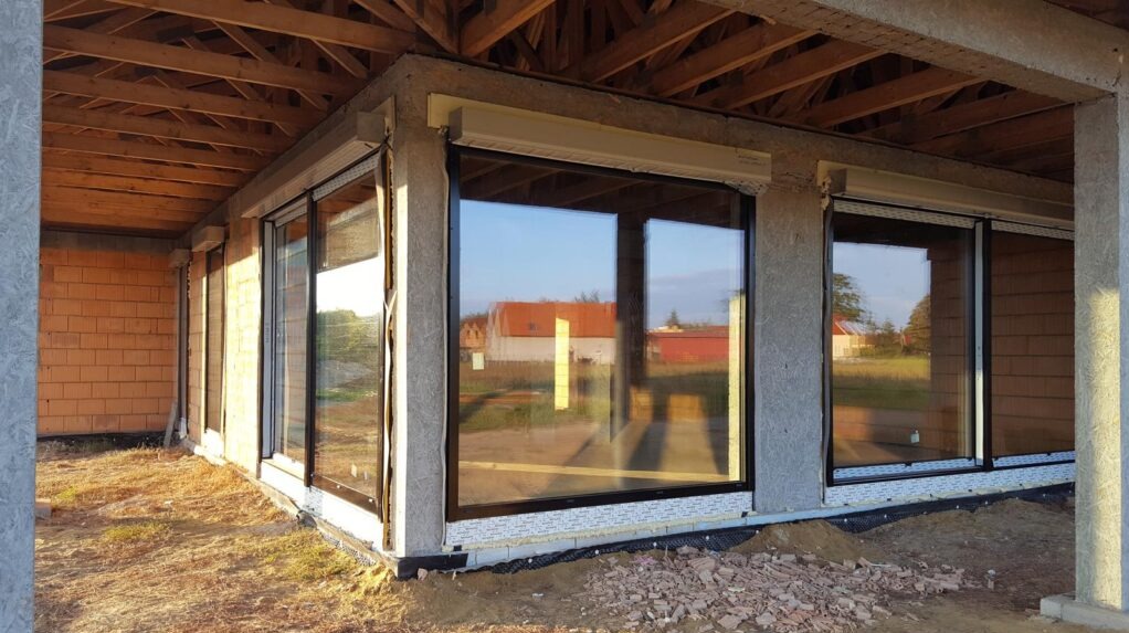 Surowy stan domu w trakcie budowy: widoczne okna z roletami zewnętrznymi, ściany z pustaków i drewniana konstrukcja dachu. W oknach odbija się krajobraz.
