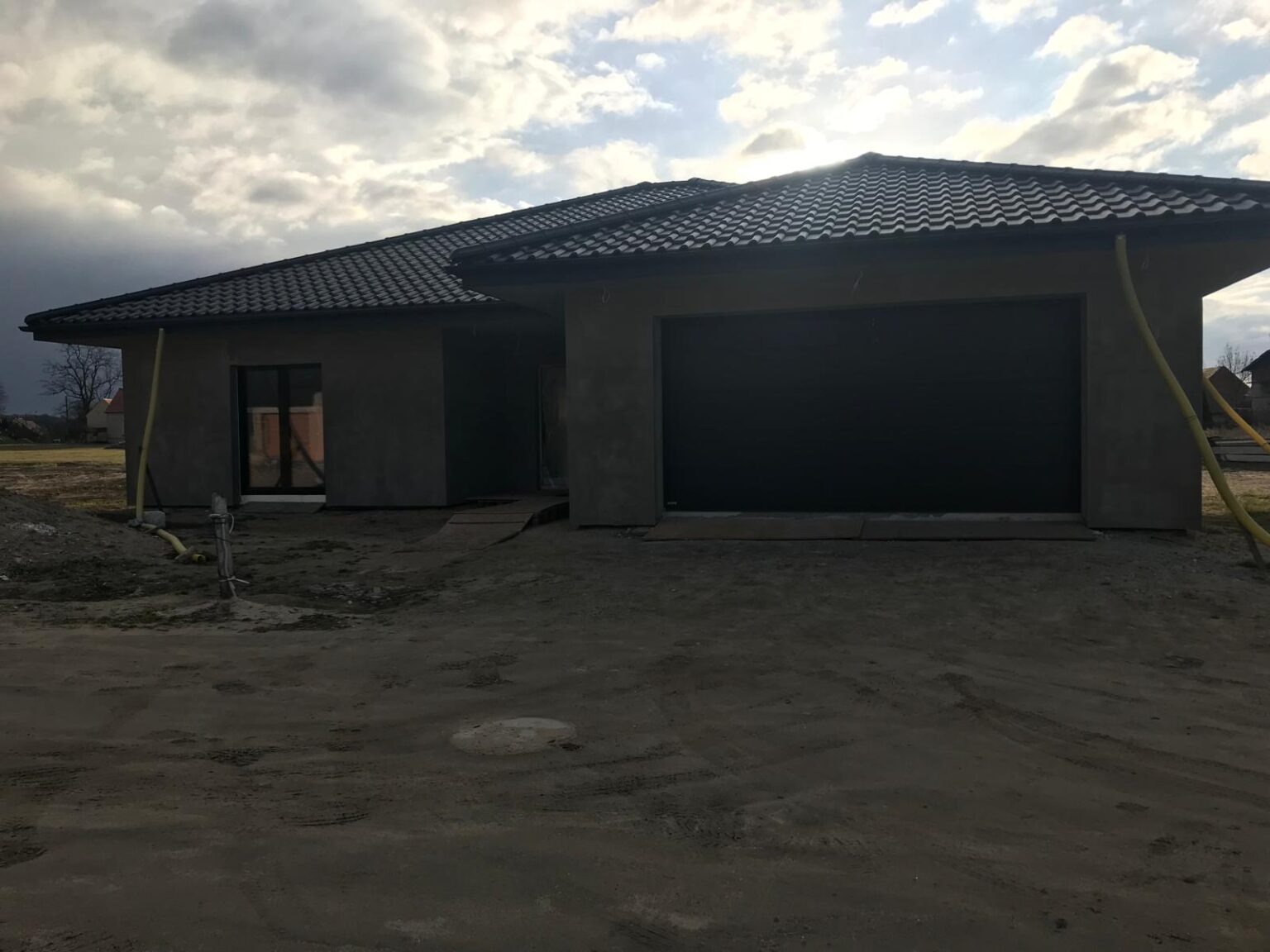 Surowy, parterowy dom z garażem w trakcie budowy, z widocznymi przewodami na elewacji i ziemią wokół, pod pochmurnym niebem.