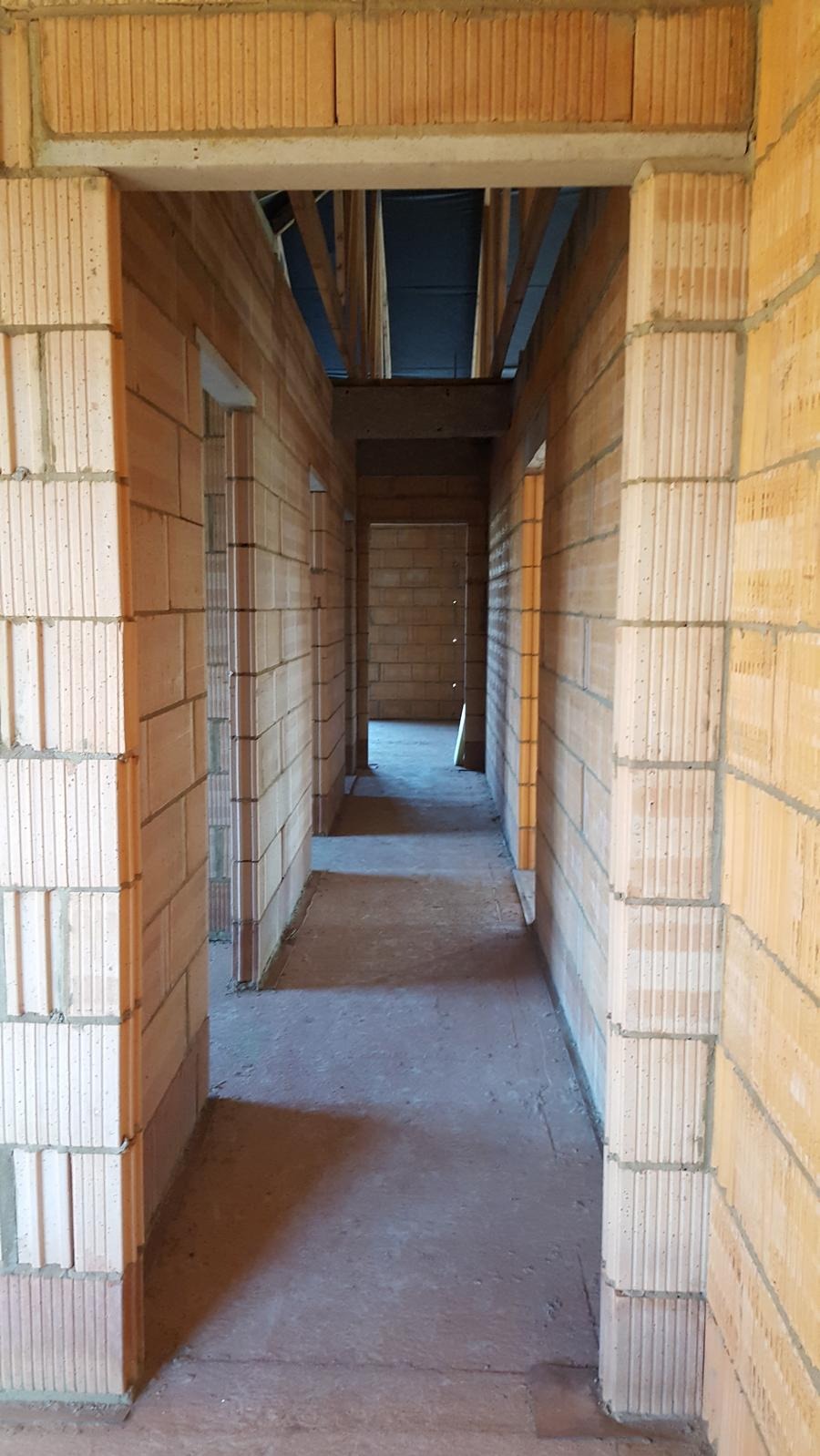 Wnętrze domu w trakcie budowy: widok korytarza z surowymi ścianami z cegieł i widoczną konstrukcją stropu. Surowy beton na podłodze. Perspektywa w głąb budynku.