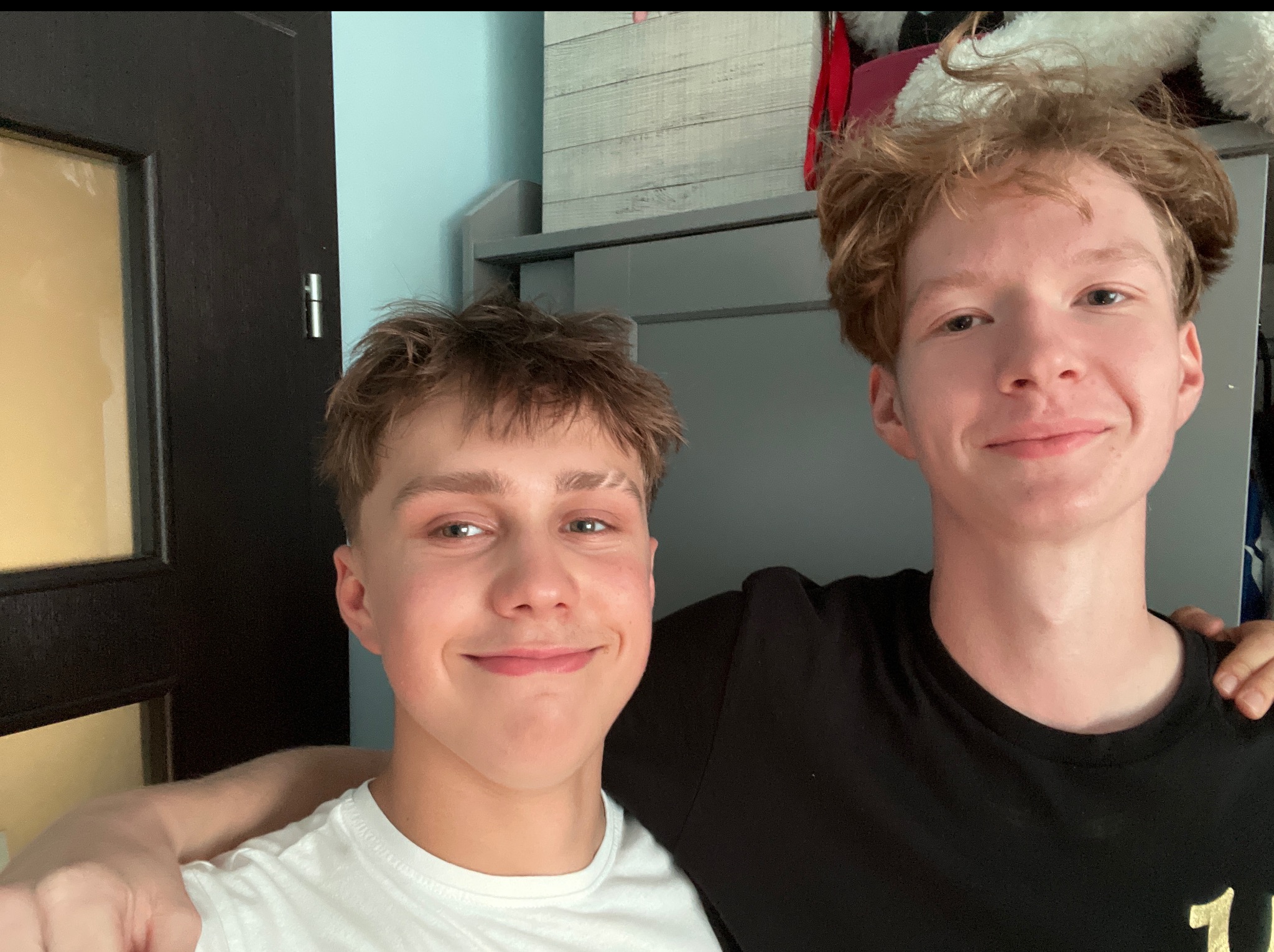 Uśmiechnięci nastolatkowie, jeden w białej koszulce, drugi w czarnej, stoją obok siebie w pokoju z błękitną ścianą i ciemnymi drzwiami. Przyjacielska atmosfera.