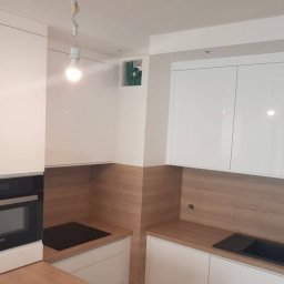 Nowoczesna zabudowa kuchenna – białe, gładkie fronty w połysku, drewniane elementy ocieplające wnętrze, podświetlenie LED oraz praktyczne rozmieszczenie szafek i sprzętów.