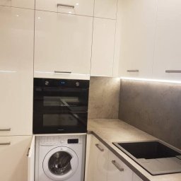 Nowoczesna zabudowa kuchenna w białym połysku – zestaw mebli z piekarnikiem, płytą indukcyjną, zmywarką i pralką w zabudowie. Szafki górne do sufitu, blat laminowany w kolorze szarości, oświetlenie LED pod szafkami.