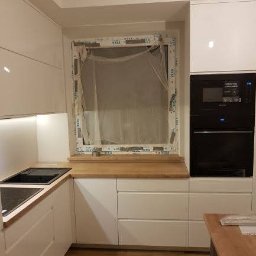 Nowoczesna zabudowa kuchenna w kształcie litery L – białe fronty w połysku, blat drewnopodobny, piekarnik i mikrofala w zabudowie, podświetlenie LED pod szafkami.