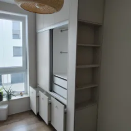 Biała szafa wnękowa z przesuwnymi drzwiami, otwarte szuflady z uchwytami haczykowymi, regał z półkami, okno z rośliną w tle, lampa sufitowa z wikliny.