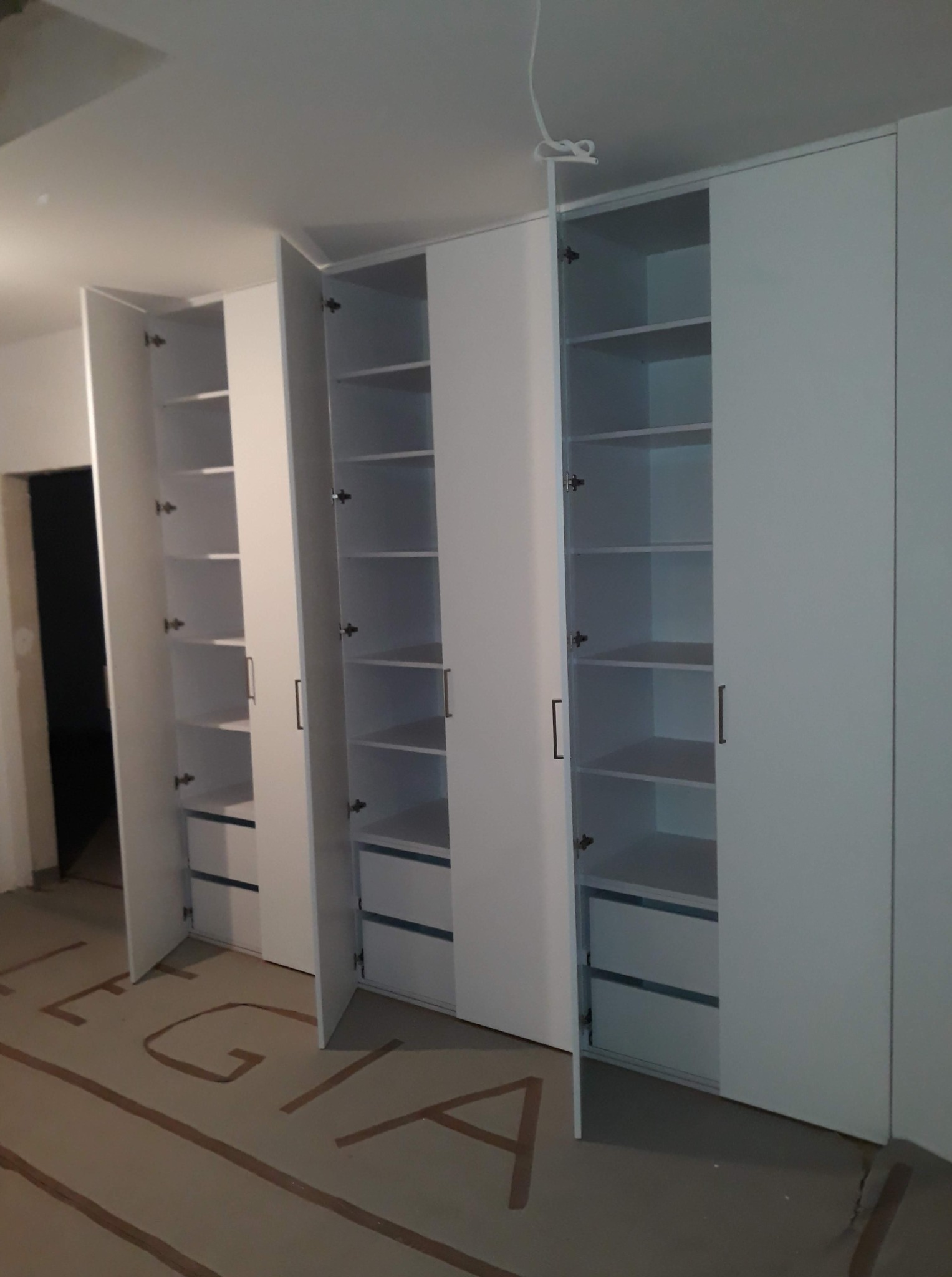 Biała, wnękowa szafa z półkami i szufladami, częściowo otwarta, widoczna na tle surowego wnętrza w trakcie wykończenia, z zabezpieczoną podłogą.