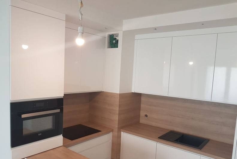 Biała kuchnia z połyskiem, drewniane akcenty, czarny piekarnik i płyta. Widoczna wentylacja w ścianie. Surowa żarówka jako oświetlenie tymczasowe.