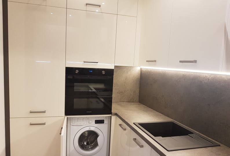 Biała, nowoczesna zabudowa kuchenna z piekarnikiem, płytą indukcyjną, zmywarką i pralką w zabudowie. Szafki do sufitu, szary blat, oświetlenie LED.