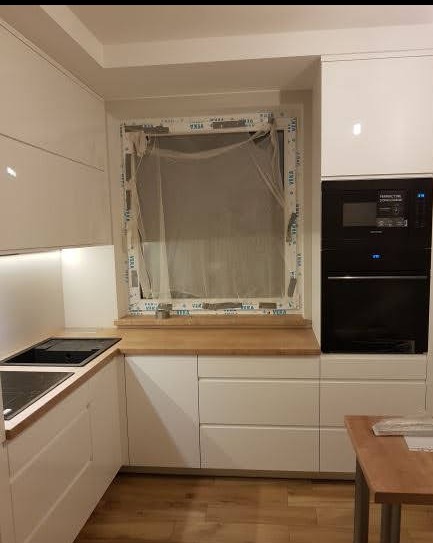 Biała kuchnia w kształcie litery L z drewnianym blatem i wbudowanym piekarnikiem. Okno z widoczną folią ochronną. Nowoczesny design, połyskujące fronty.