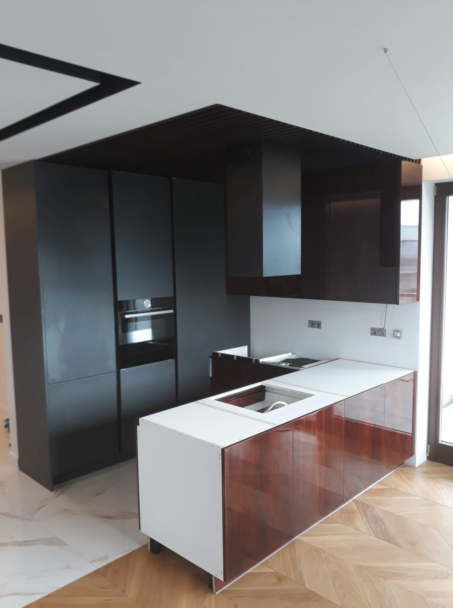 Nowoczesna kuchnia z wyspą – grafitowe fronty, biały blat z drewnem, na suficie panele ażurowe (słabo widoczne).