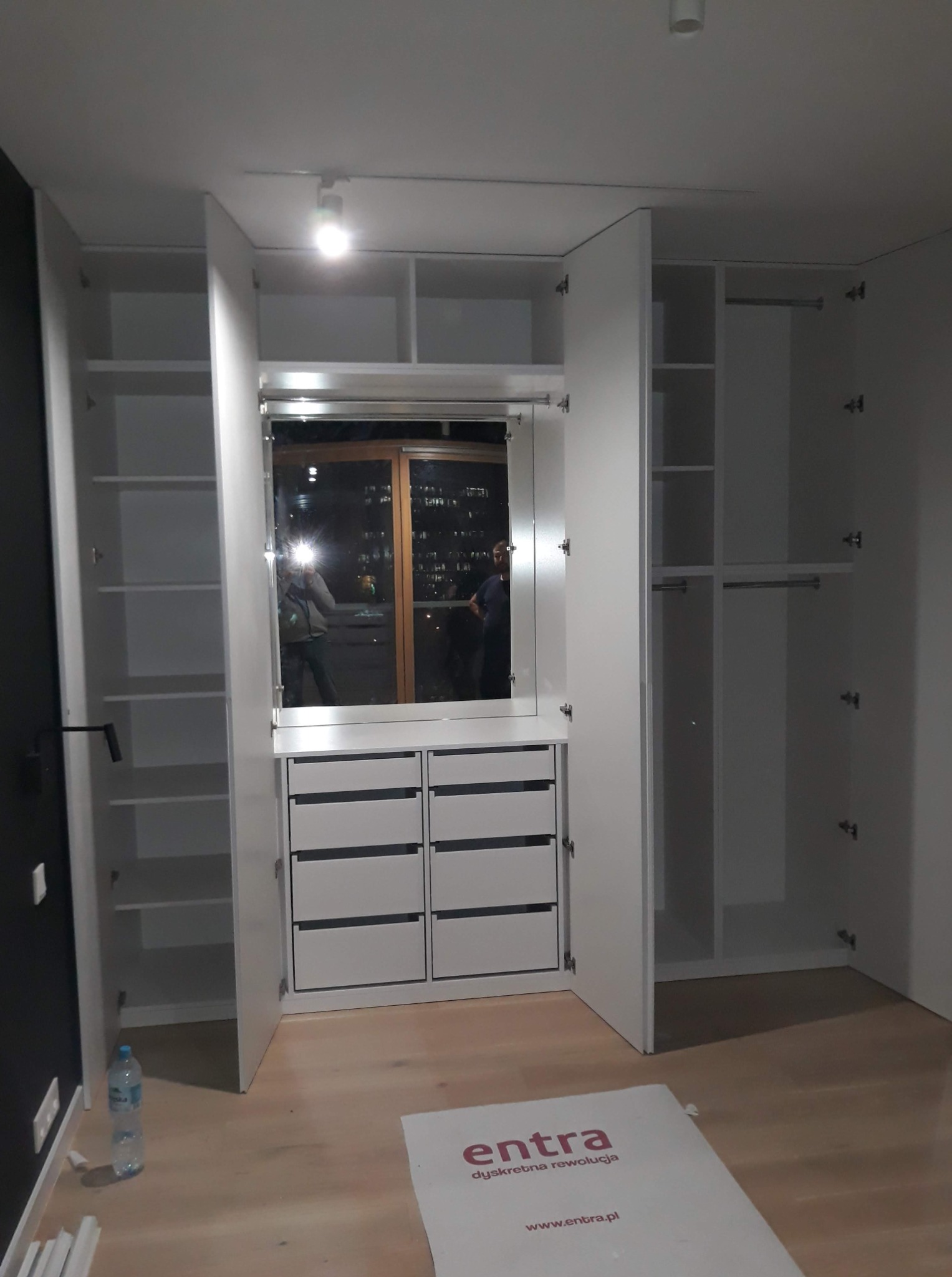 Biała szafa wnękowa z lustrem i szufladami, otwarte drzwi ukazują półki i drążki. Na podłodze leży karton. Widoczne odbicie osoby w lustrze.