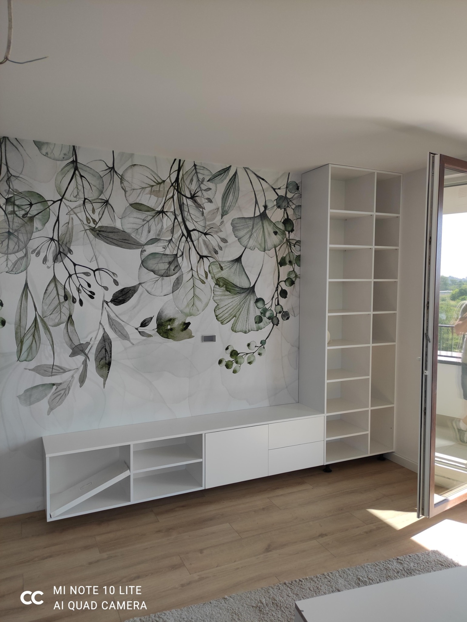 Zabudowa RTV w bieli z ażurową półką i dekoracyjną ścianą z grafiką roślinną.