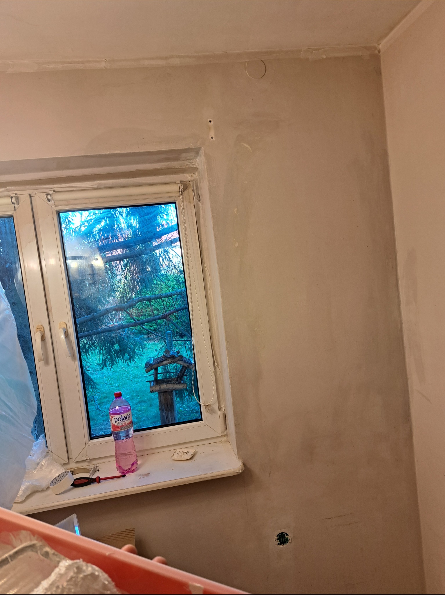 Ściana po szpachlowaniu, widoczne ślady po łatach, okno z widokiem na zewnątrz, butelka wody i narzędzia na parapecie, przygotowanie do malowania.