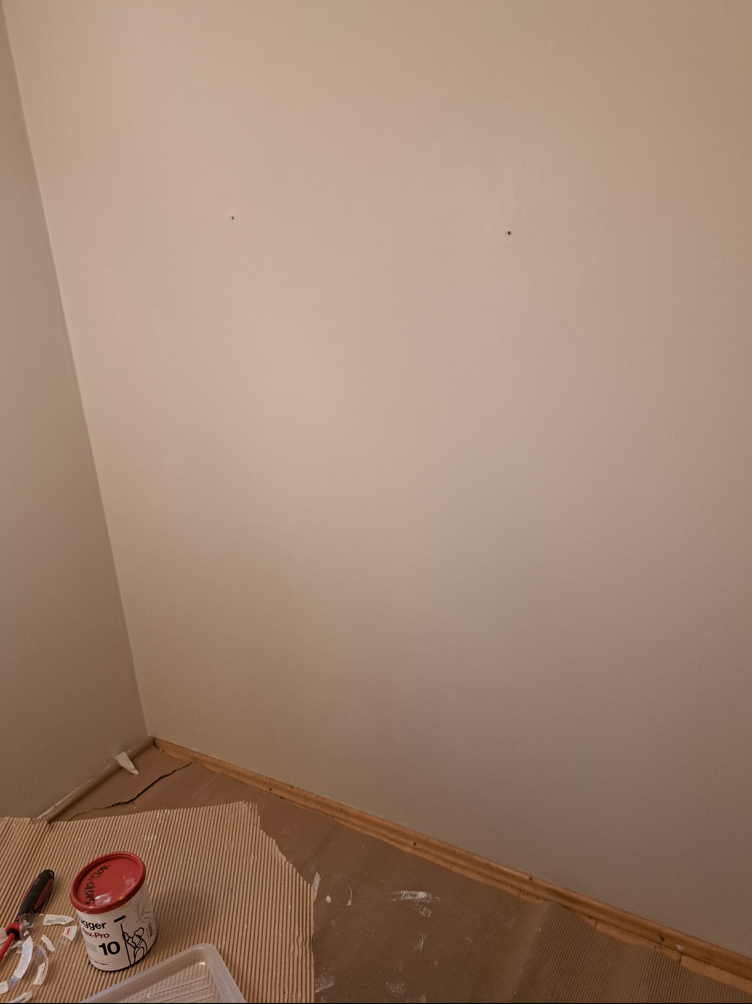 Ściana w trakcie malowania, widoczne dwie dziury po kołkach, zabezpieczona podłoga, puszka farby i narzędzia malarskie w rogu pomieszczenia.