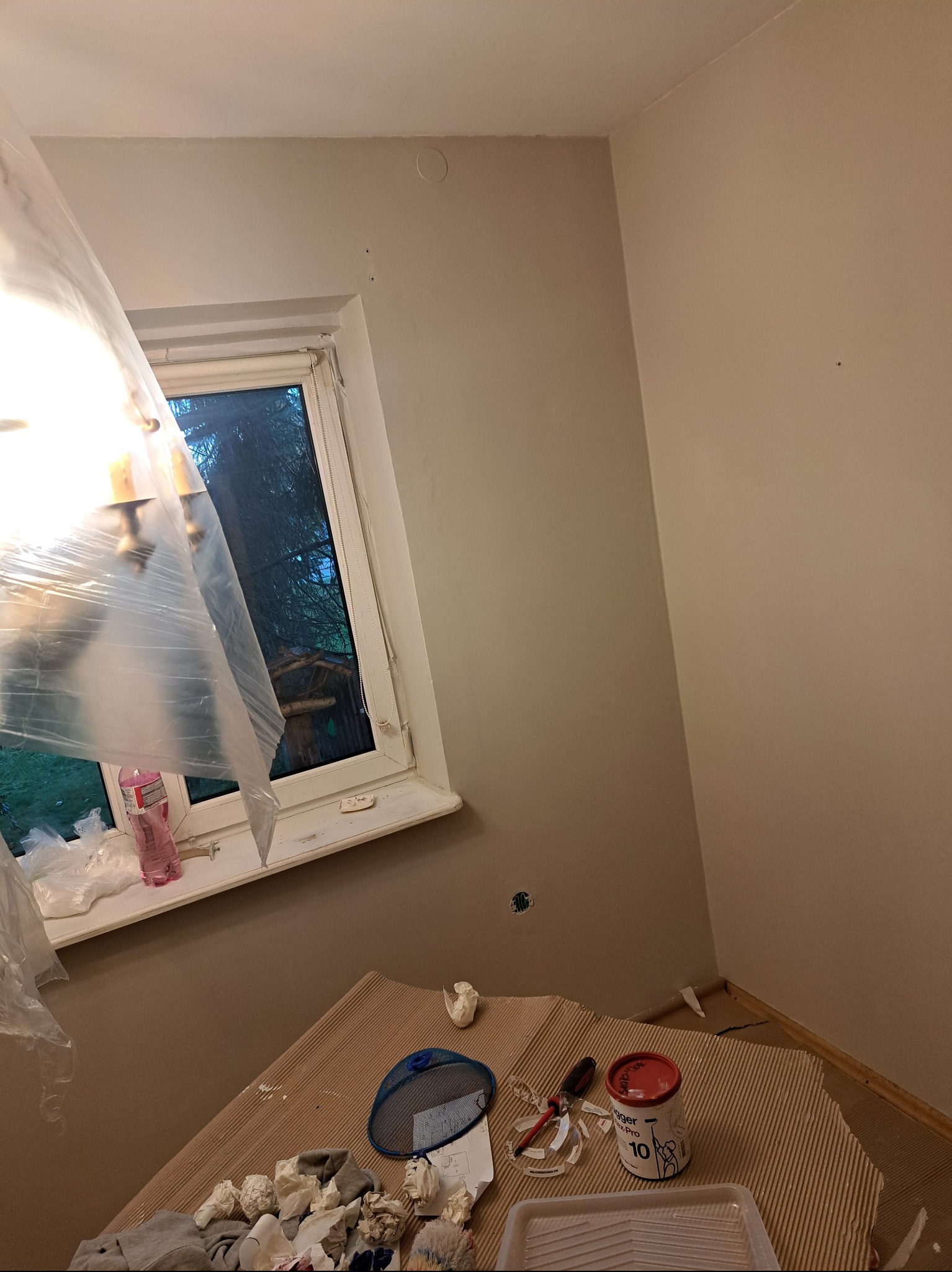 Pokój w trakcie odświeżania: pomalowane ściany w odcieniach beżu, okno z widokiem na zewnątrz, zabezpieczona lampa, farba i narzędzia malarskie na stole.
