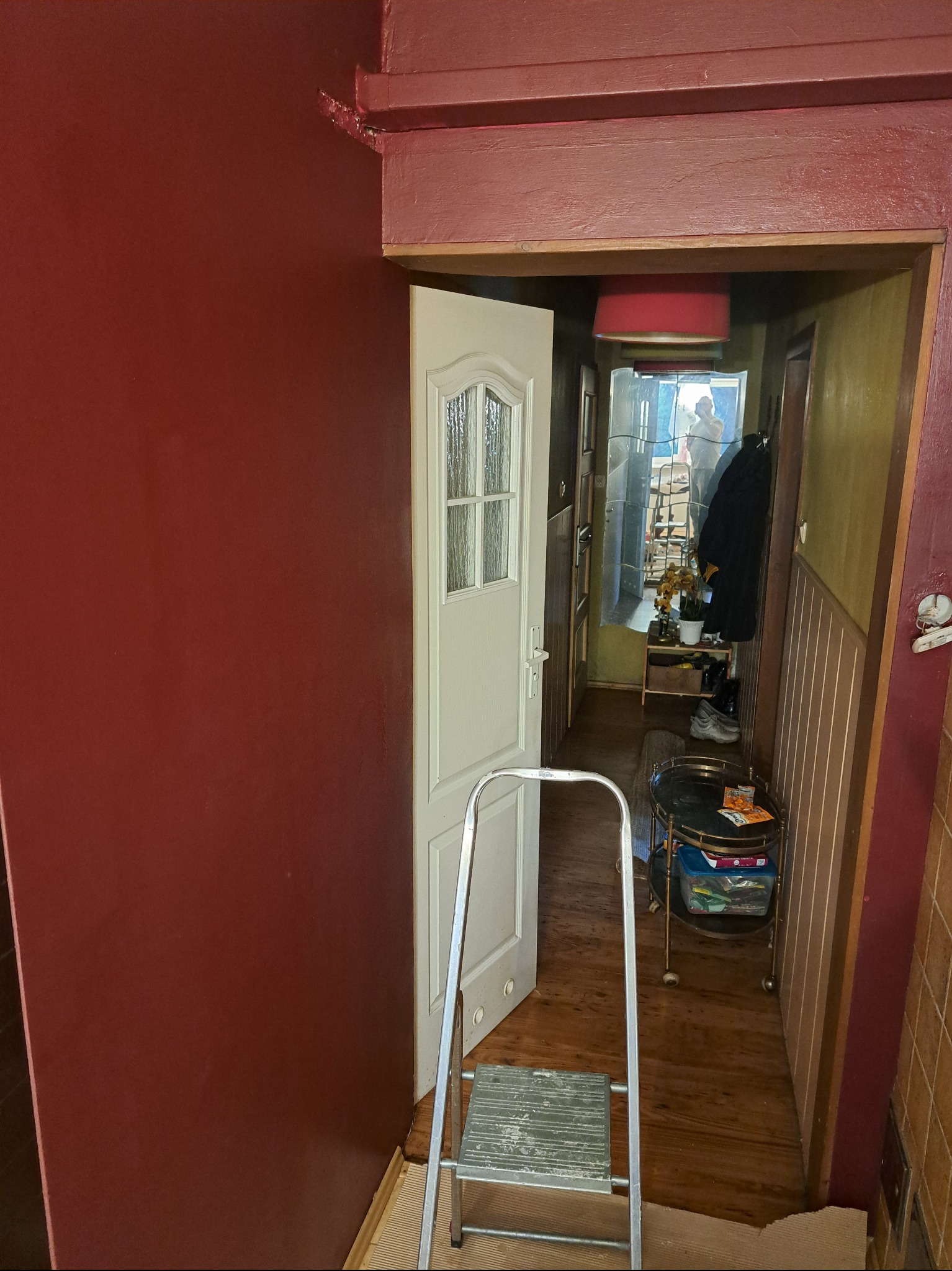 Ściana w odcieniach czerwieni po odświeżeniu, widoczna drabina malarska i otwarte drzwi do przedpokoju. Prace wykończeniowe w trakcie realizacji.