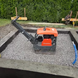 Zagęszczarka Husqvarna ubija żwir w fundamencie w Pszczynie. Izolacja pionowa fundamentu z naniesioną warstwą hydroizolacyjną. Widoczne szalunki.