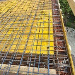 KAMIL TARGONI USŁUGI OGÓLNOBUDOWLANE I STOLARSKIE - Żelbetowa konstrukcja stropu z żółtym szalunkiem i zbrojeniem z prętów stalowych, widok z góry, stopa w bucie roboczym w rogu kadru.