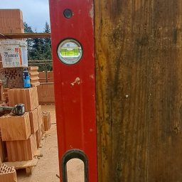 KAMIL TARGONI USŁUGI OGÓLNOBUDOWLANE I STOLARSKIE - Czerwona poziomica przyłożona do drewnianej belki, w tle paleta z cegłami i wiadro farby. Sprawdzanie pionu konstrukcji.