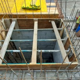 KAMIL TARGONI USŁUGI OGÓLNOBUDOWLANE I STOLARSKIE - Szczyt ujęcia szalunku zbrojonego na fundamencie domu z cegły, z pracownikiem w kasku i kamizelce mierzącym ścianę. Widok z góry na konstrukcję.