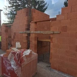 KAMIL TARGONI USŁUGI OGÓLNOBUDOWLANE I STOLARSKIE - Wznoszenie ścian z czerwonej cegły Wienerberger na budowie. Widoczne zbrojenia i drewniane podpory. Częściowo zapakowane cegły na pierwszym planie.