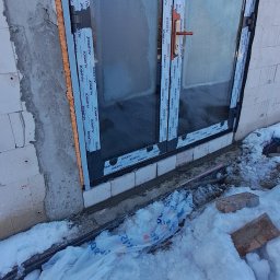 KAMIL TARGONI USŁUGI OGÓLNOBUDOWLANE I STOLARSKIE - Montaż okna Alufrof w surowym murze, widoczne ocieplenie pianką montażową i śnieg na zewnątrz. Okno z szybą pokrytą szronem.