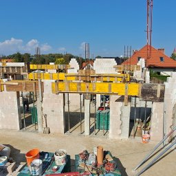 KAMIL TARGONI USŁUGI OGÓLNOBUDOWLANE I STOLARSKIE - Budowa domu jednorodzinnego: mury z bloczków, szalunki drewniane, zbrojenia, dźwig w tle. Widok z góry na plac budowy w słoneczny dzień.