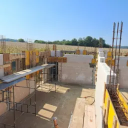 Budowa domu jednorodzinnego: fundamenty z bloczków betonowych, szalunki i zbrojenia na tle pola kukurydzy w słoneczny dzień. Etap wznoszenia ścian.