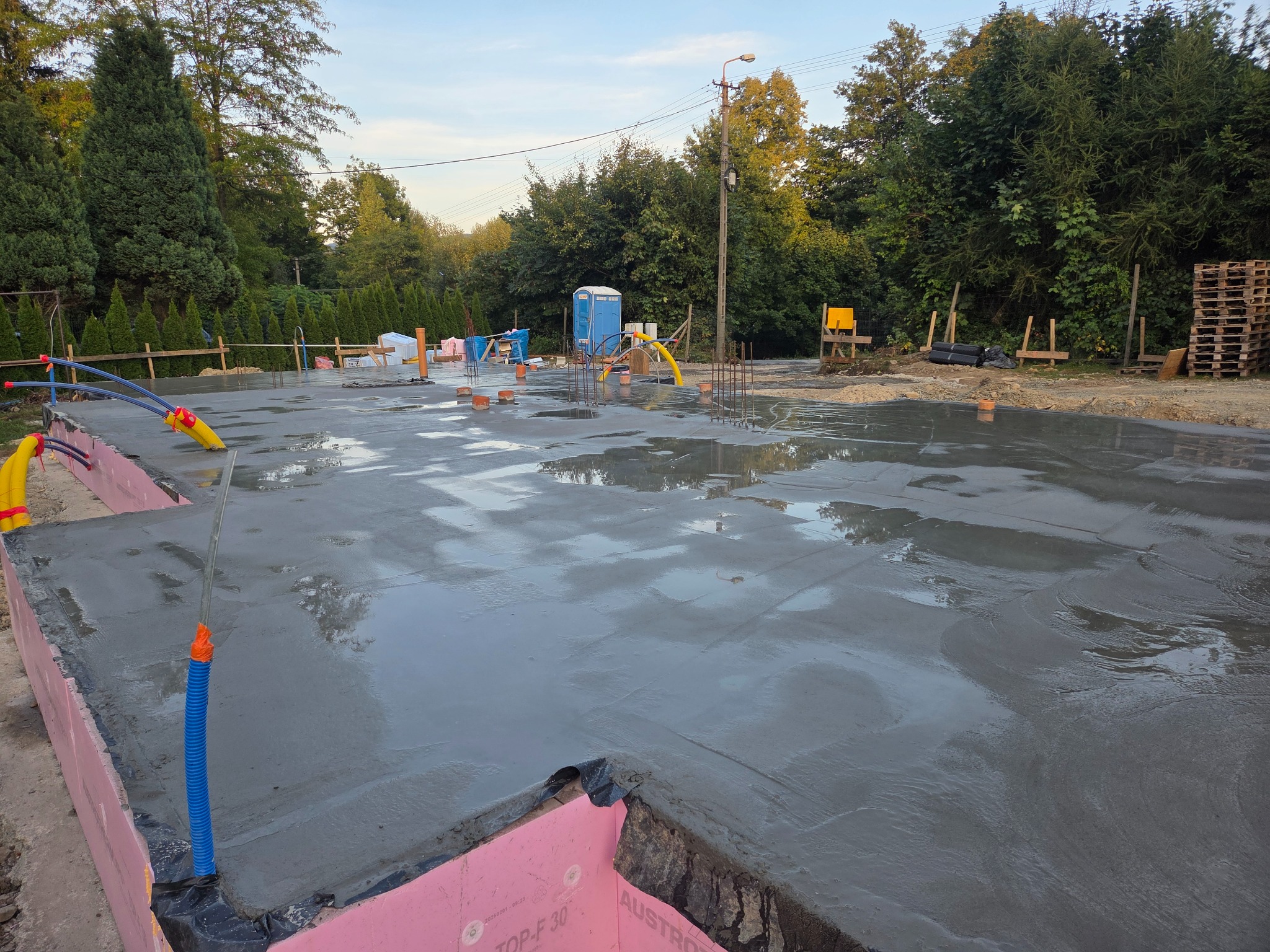 Zalany fundament pod budynek z widoczną izolacją termiczną i wystającymi prętami zbrojeniowymi. W tle drzewa i przenośna toaleta.