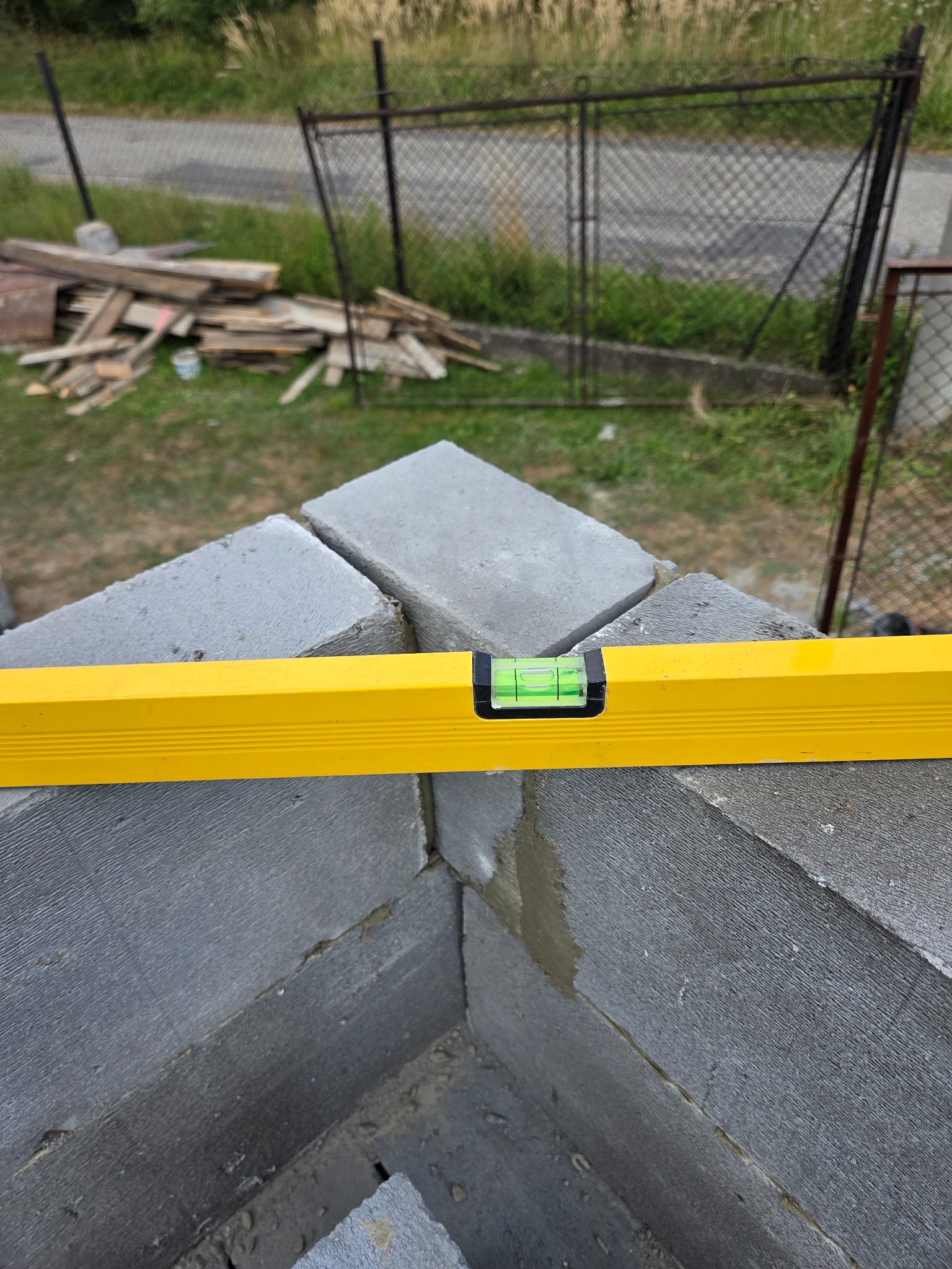 Wyrównywanie bloczków betonowych żółtą poziomicą w trakcie budowy ściany. Widoczny fragment muru, w tle teren budowy z elementami ogrodzenia i składowanym drewnem.