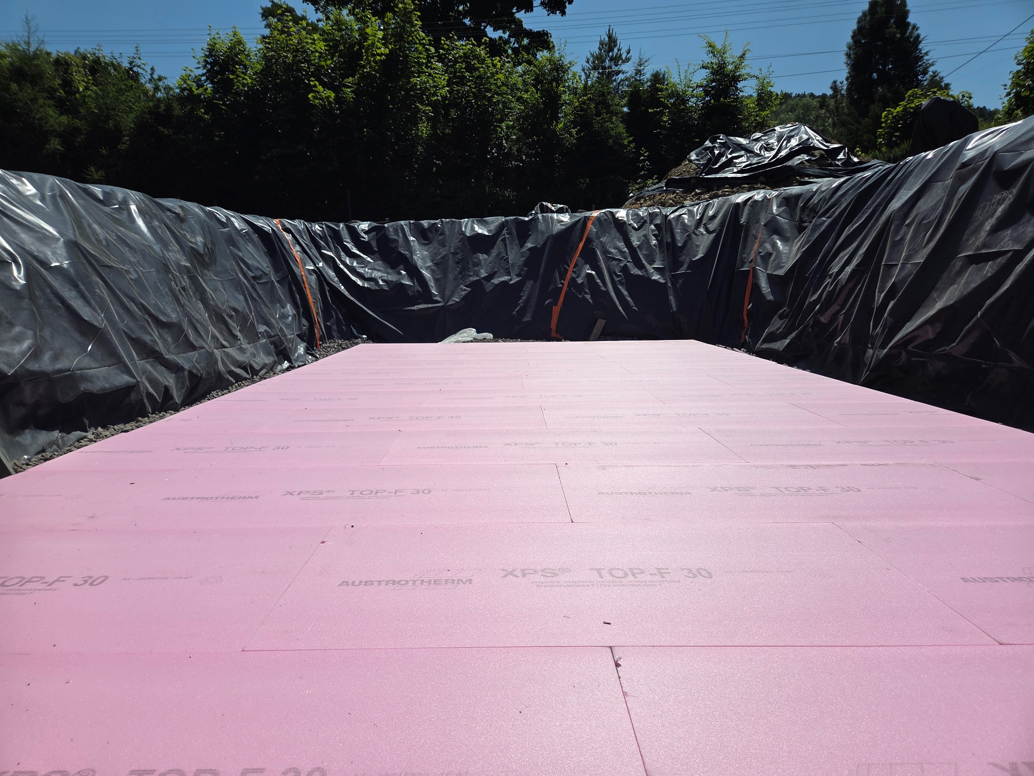 Wyłożony różowy styropian Austrotherm XPS TOP-F 30 na dnie wykopu zabezpieczonego czarną folią, przygotowanego pod fundamenty budynku na tle zieleni.