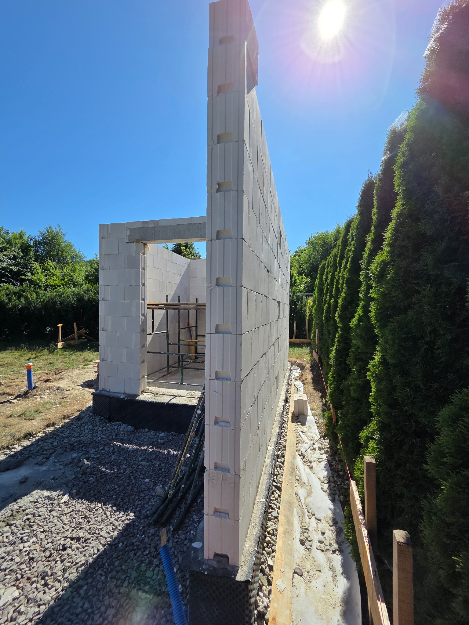 Wznoszenie ścian parterowego domu z białych bloczków, widoczne rusztowanie wewnątrz, tło: zielone tuje i błękitne niebo. Budowa w słoneczny dzień.