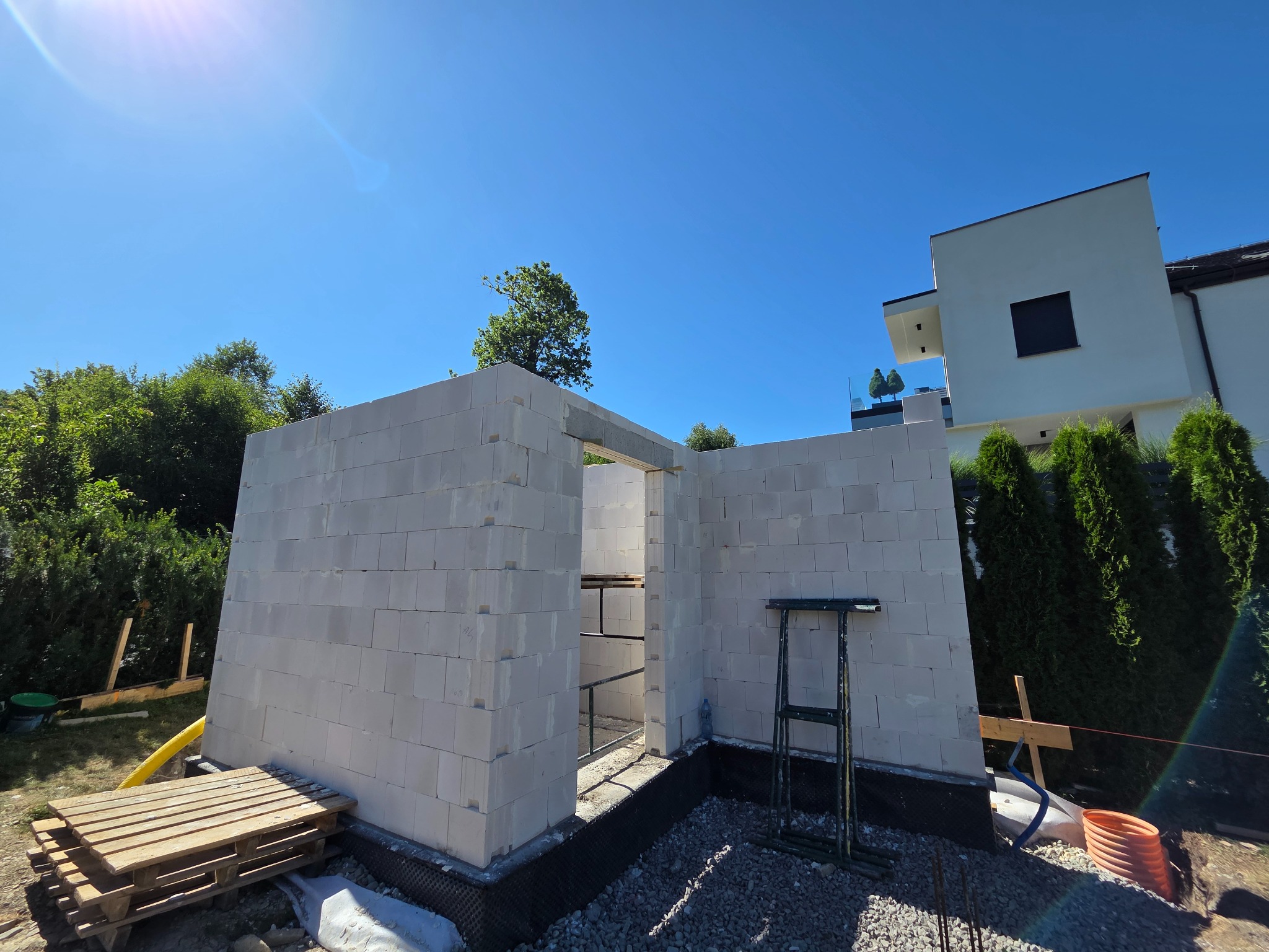 Wznoszenie ścian z bloczków betonu komórkowego Ytong na fundamencie. Widoczne zbrojenie, paleta z materiałami i fragment budynku w tle w słoneczny dzień.