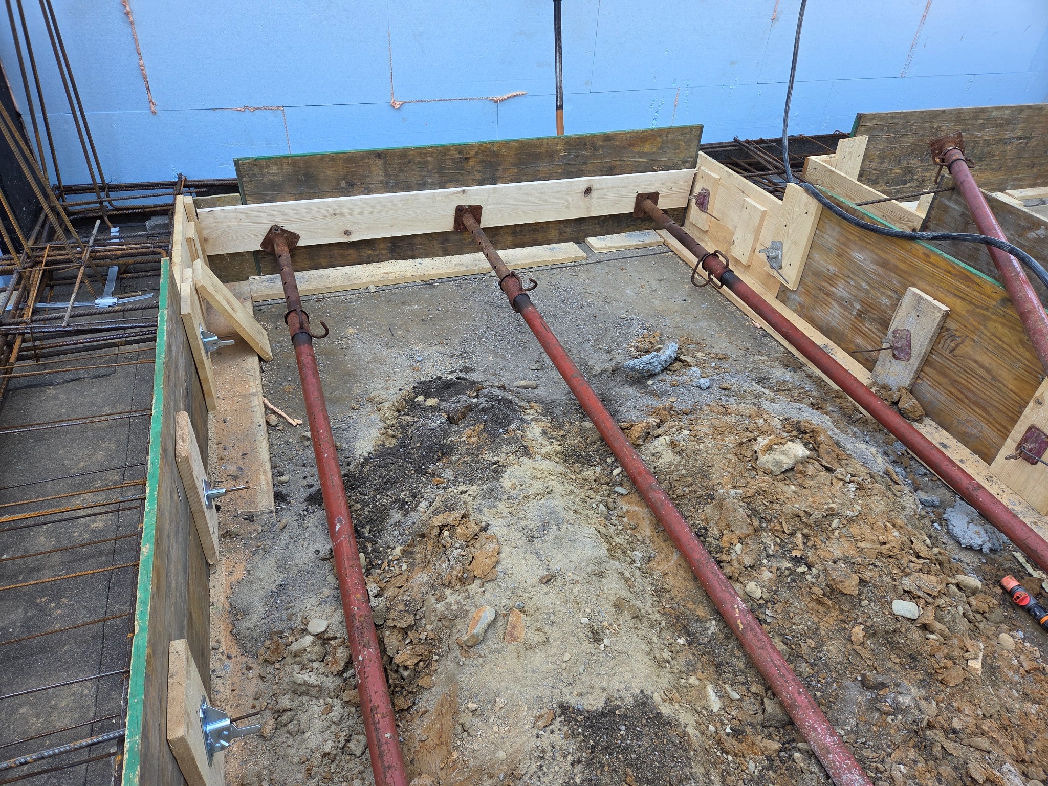 Szczegółowe ujęcie szalunku fundamentów z drewnianymi deskami i metalowymi podporami, ukazujące zbrojenie i przygotowanie do wylania betonu.