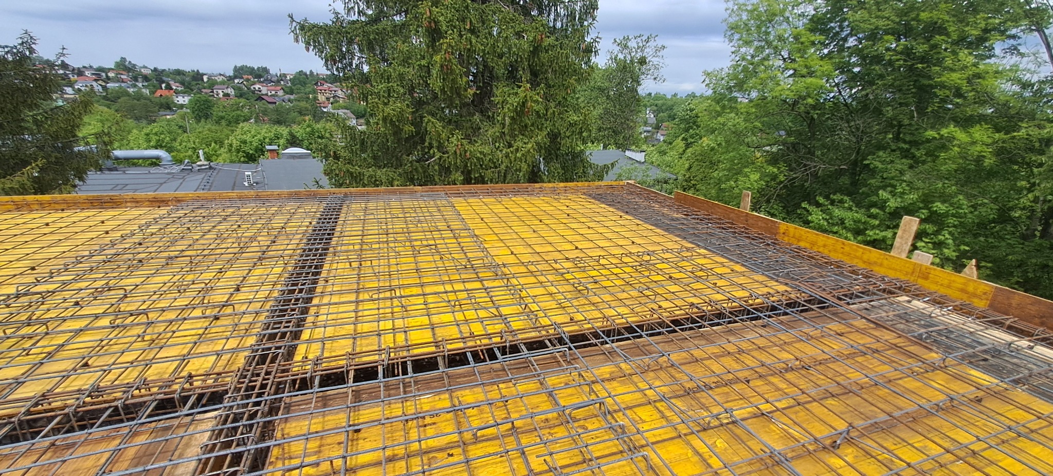 Żółte szalunki i zbrojenie stropu na tle zieleni i zabudowy miejskiej. Widok z góry, etap budowy domu.
