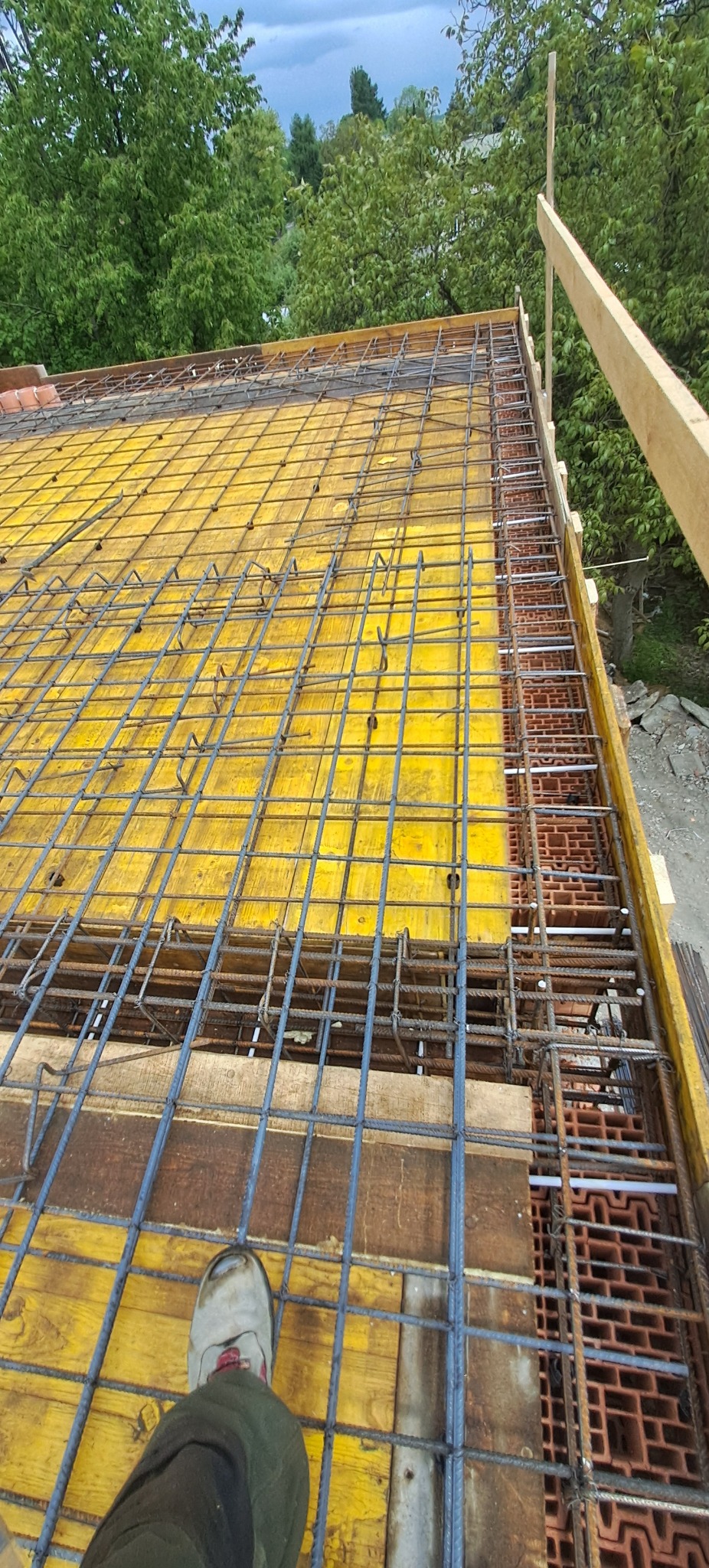 Żelbetowa konstrukcja stropu z żółtym szalunkiem i zbrojeniem z prętów stalowych, widok z góry, stopa w bucie roboczym w rogu kadru.