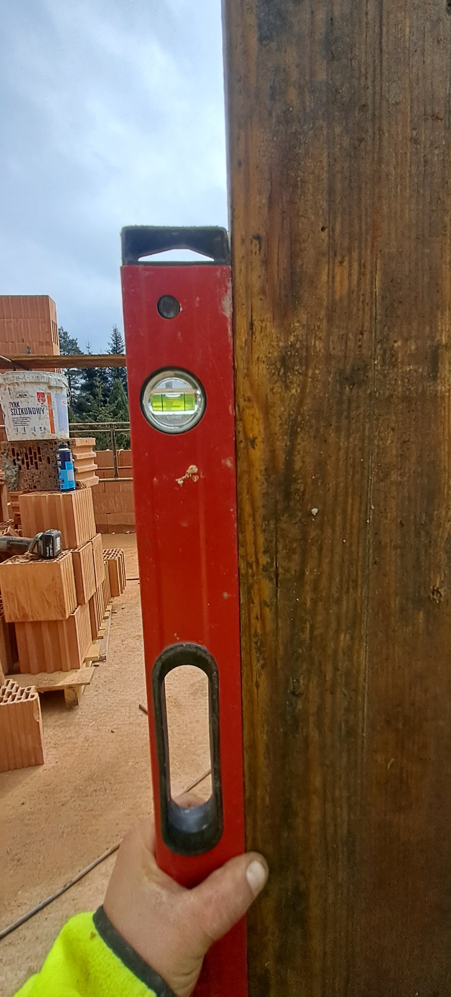 Czerwona poziomica przyłożona do drewnianej belki, w tle paleta z cegłami i wiadro farby. Sprawdzanie pionu konstrukcji.