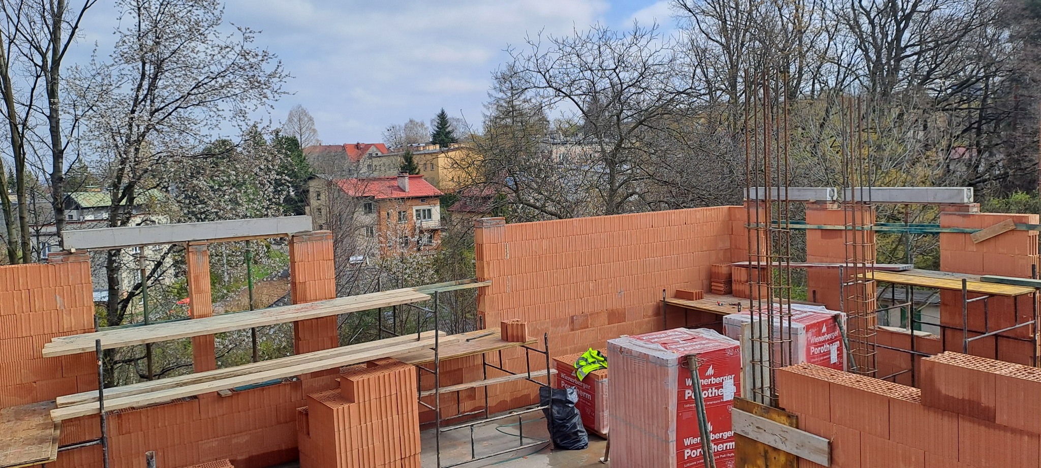 Wznoszenie murów z cegły Porotherm, widoczne zbrojenia i elementy konstrukcyjne. W tle zabudowa miejska z kwitnącymi drzewami.