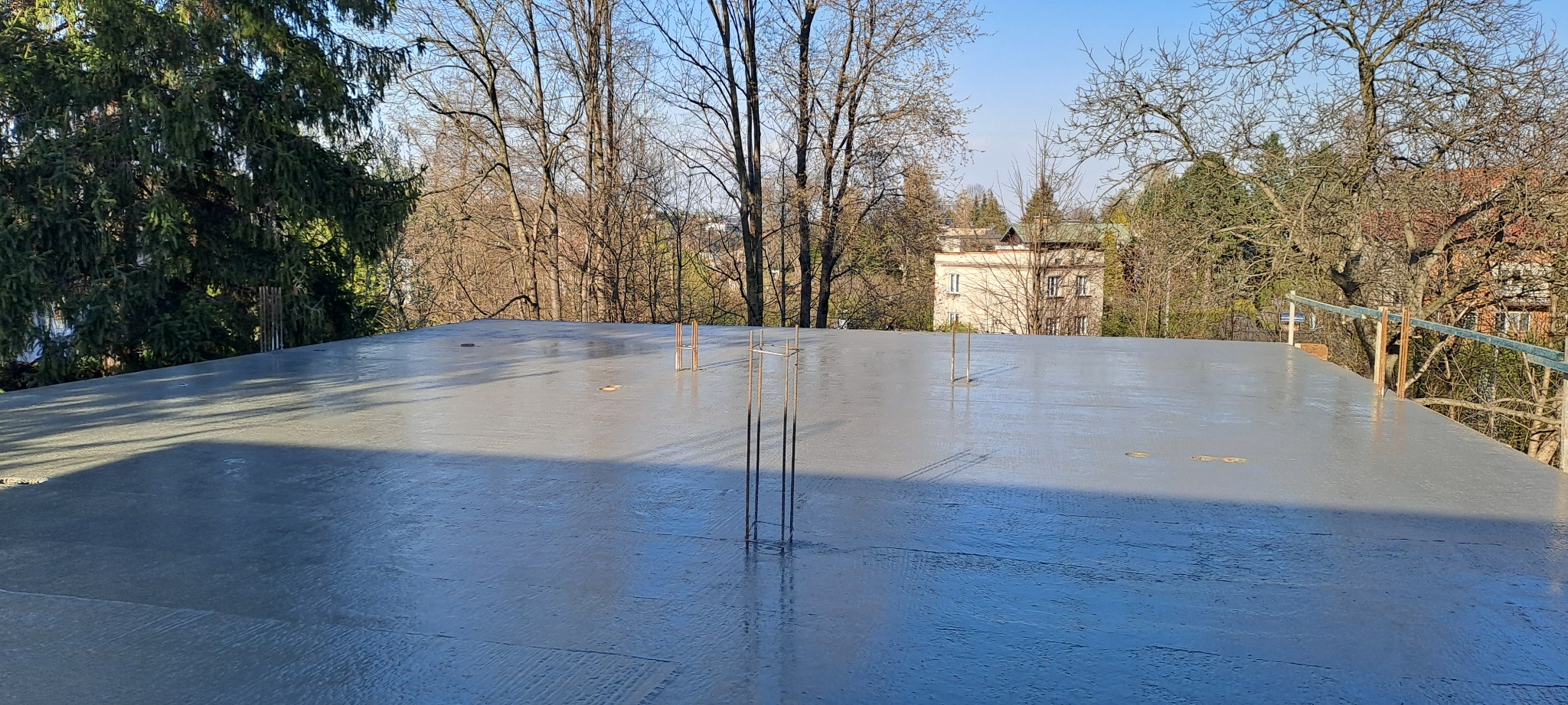 Wylany, świeży strop żelbetowy z wystającymi prętami zbrojeniowymi. W tle drzewa i budynek w oddali, błękitne niebo. Widok z góry na plac budowy.