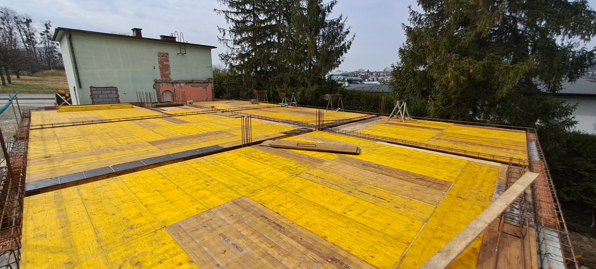Żółte szalunki stropowe zbrojone stalą na budowie domu, widok z góry na tle budynku i drzew. Etap przygotowania do zalewania betonem.