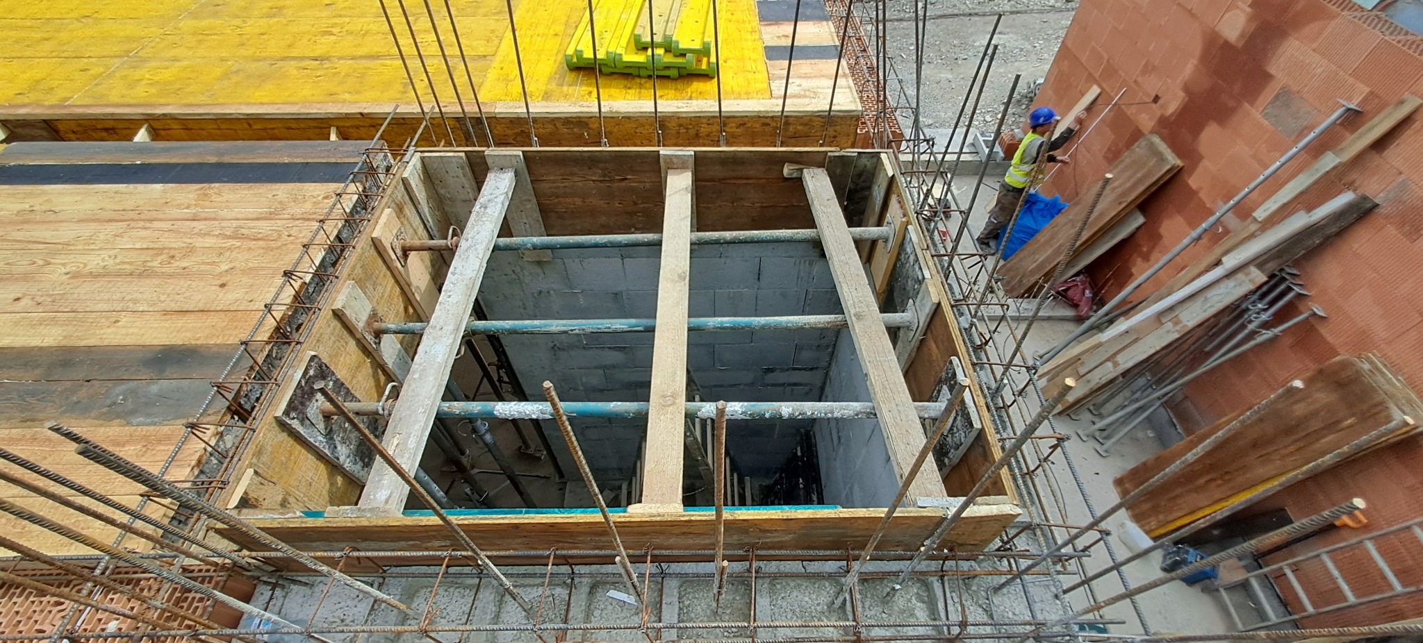 Szczyt ujęcia szalunku zbrojonego na fundamencie domu z cegły, z pracownikiem w kasku i kamizelce mierzącym ścianę. Widok z góry na konstrukcję.