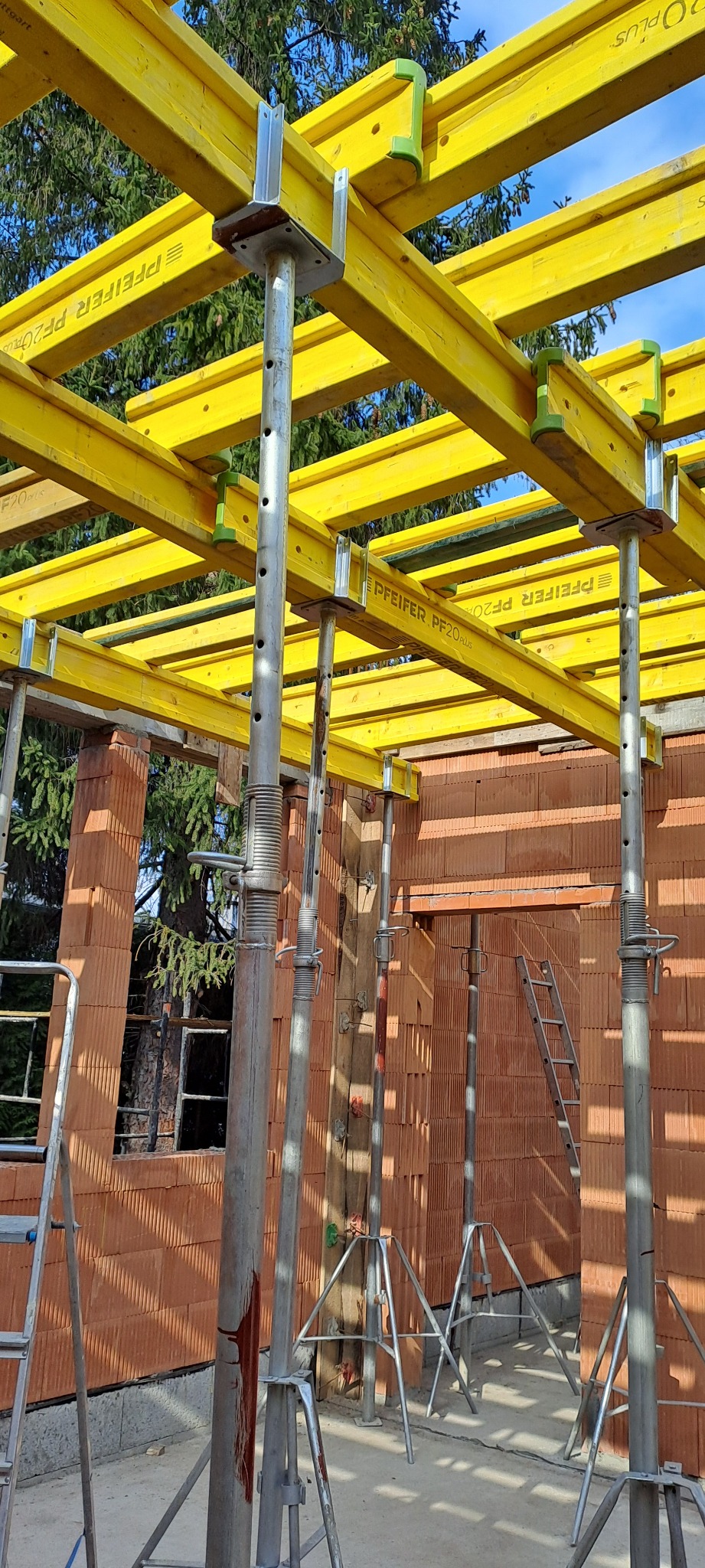 Wznoszenie murów z pustaków ceramicznych. Żółte, drewniane stemple podtrzymują strop. Widoczne drabiny i elementy szalunków stropowych.