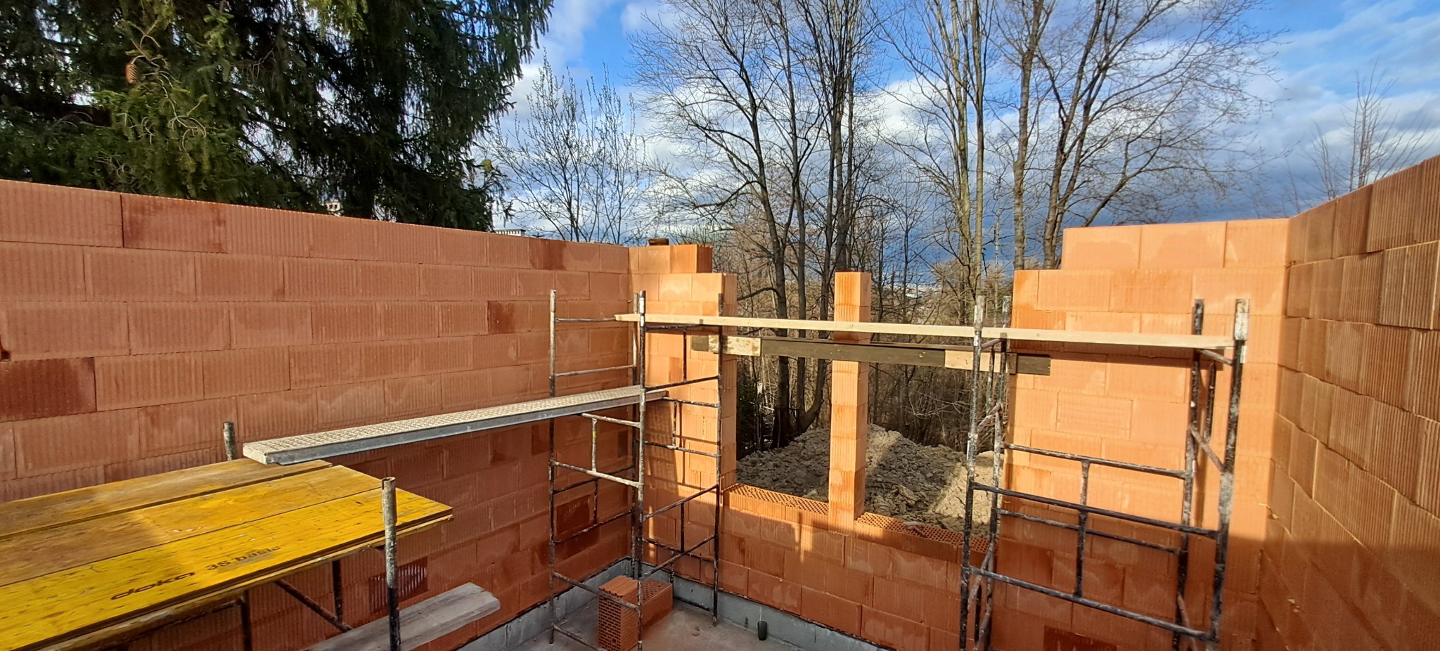 Wznoszenie muru z czerwonej cegły ceramicznej, widoczne rusztowania i fragmenty drewnianych belek. W tle drzewa i błękitne niebo.