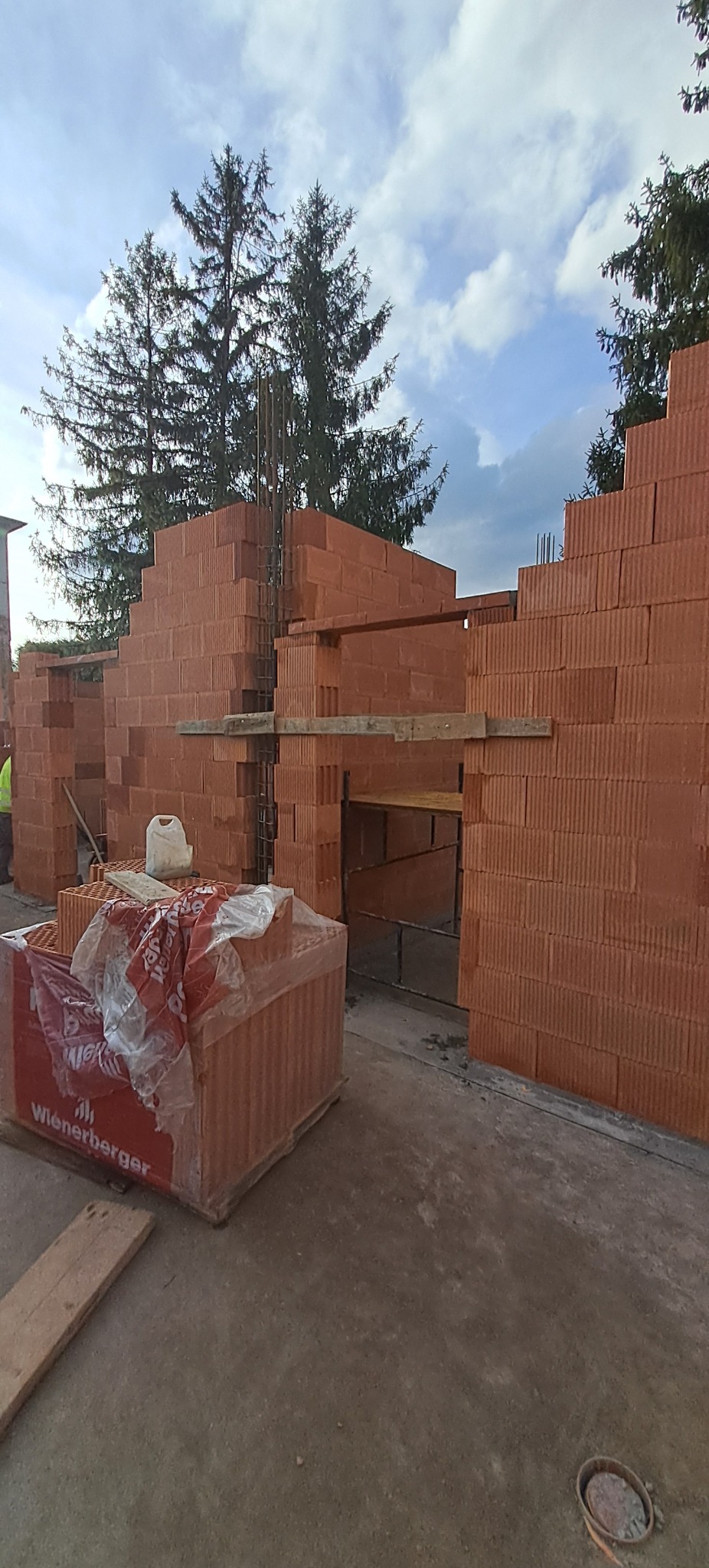 Wznoszenie ścian z czerwonej cegły Wienerberger na budowie. Widoczne zbrojenia i drewniane podpory. Częściowo zapakowane cegły na pierwszym planie.