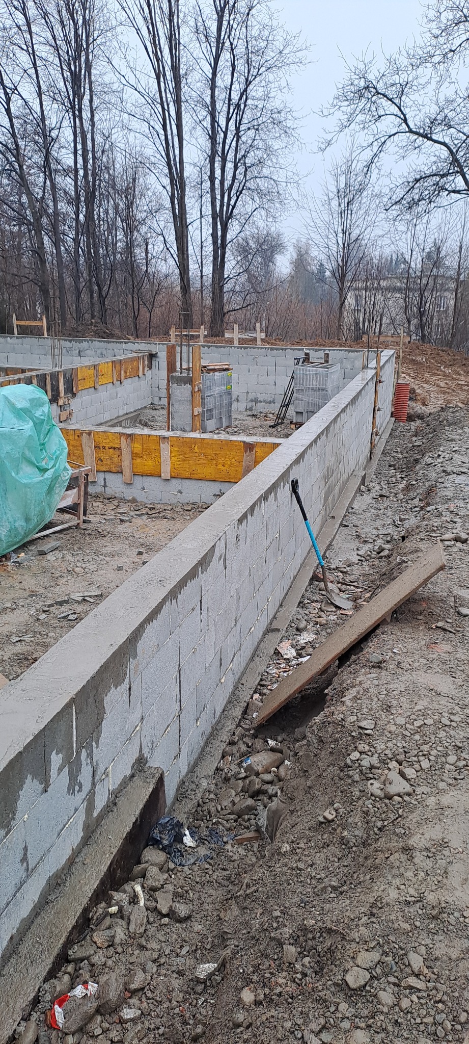 Fundamenty domu w trakcie budowy, widoczne bloczki betonowe, szalunki i wykop. Na pierwszym planie łopata wbita w ziemię. Drzewa w tle.