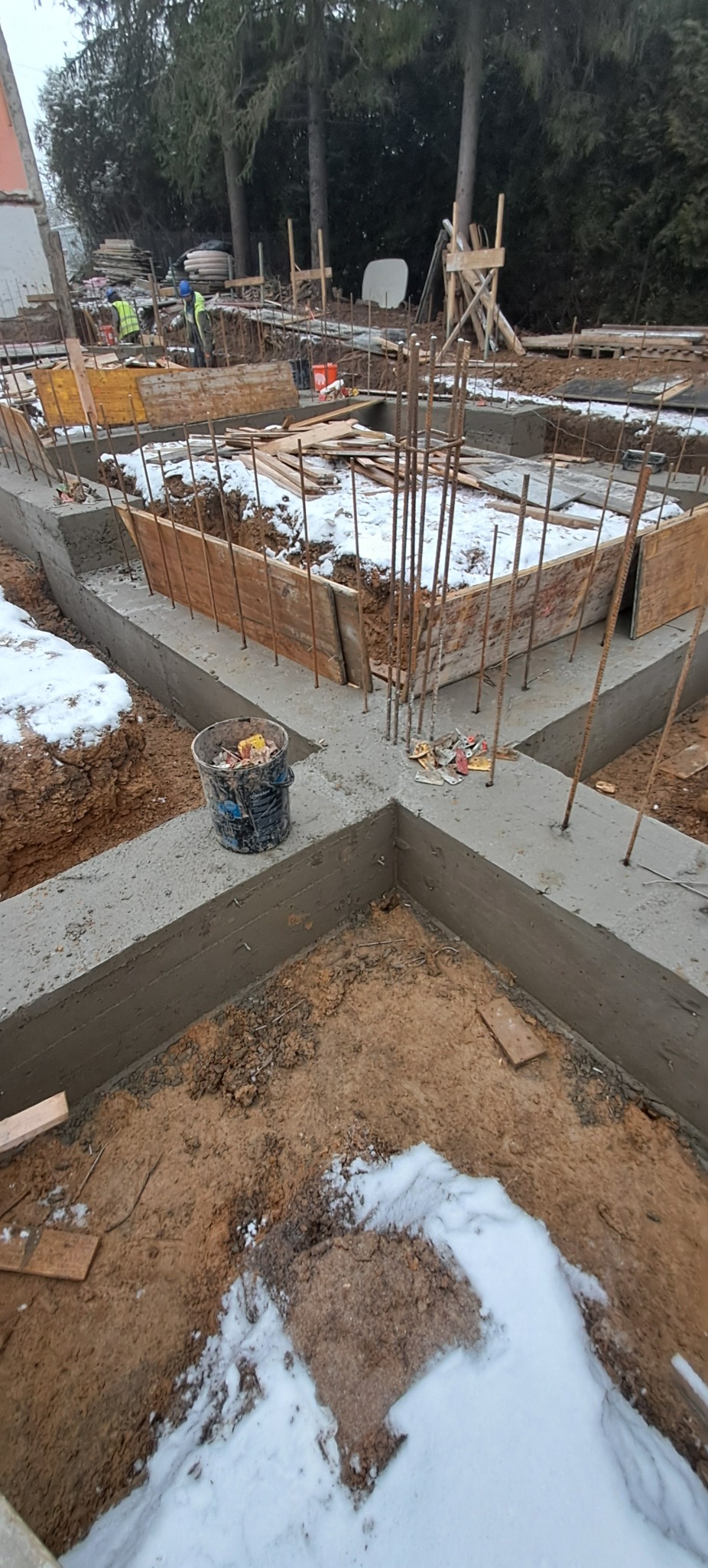 Zalane fundamenty pod budowę domu, z widocznym zbrojeniem i deskowaniem. W tle drzewa i częściowo widoczny budynek w zimowej scenerii.