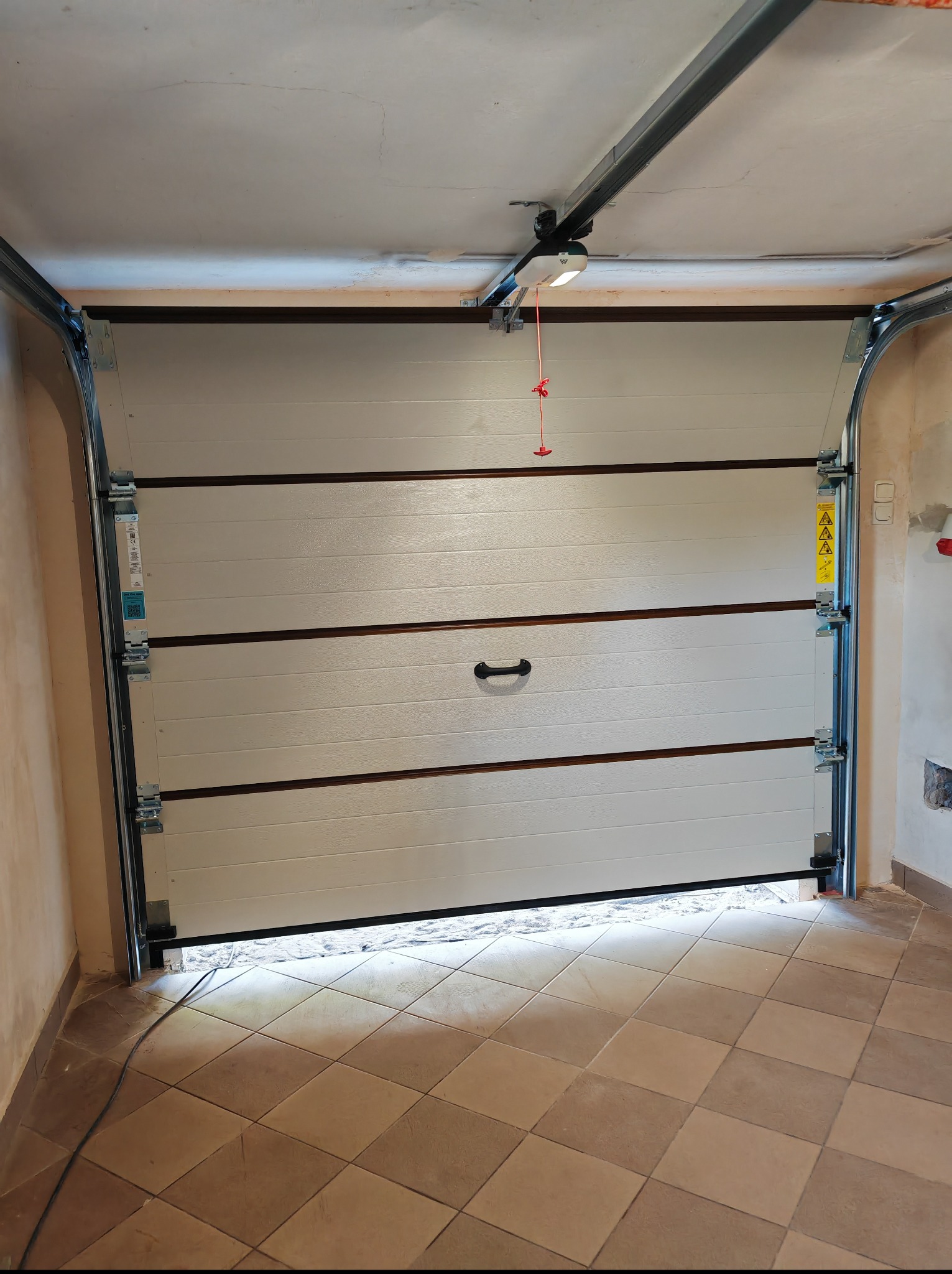 Otwarta segmentowa brama garażowa w kolorze kremowym, z widocznym mechanizmem automatycznym na suficie garażu i częściowo widocznymi płytkami podłogowymi.
